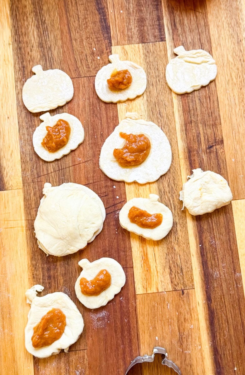 Pumpkin-shaped puff pastry cutouts topped with homemade pumpkin pie filling on a wooden cutting board, ready to assemble into mini pumpkin pie hand pies.