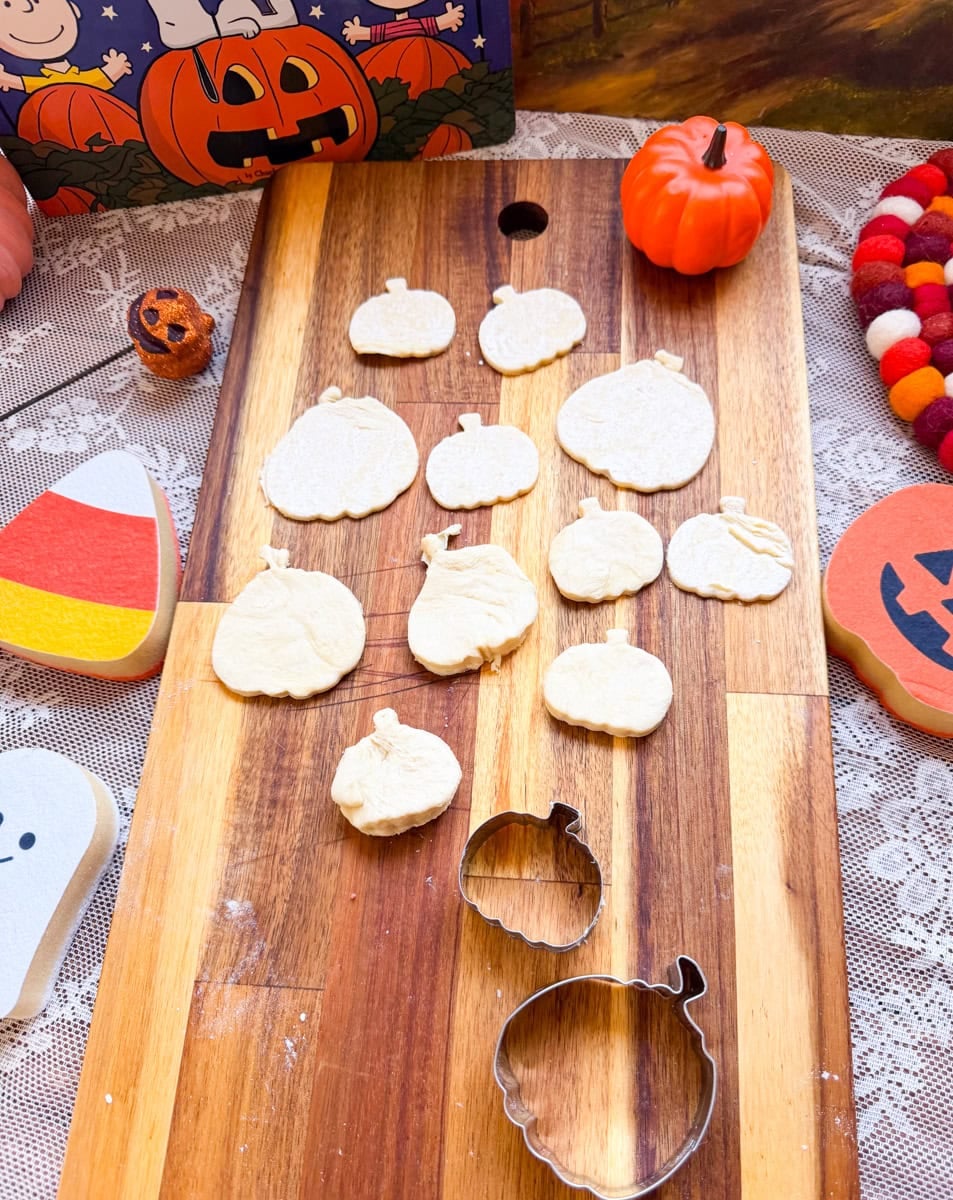 Pumpkin-shaped puff pastry cutouts arranged on a wooden cutting board with Halloween pumpkin cookie cutters, ready to be filled with homemade pumpkin pie filling.