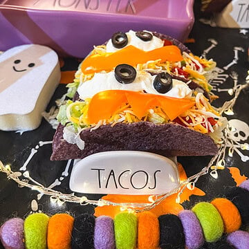 Two Halloween monster tacos in purple shells decorated with sour cream eyes, black olives, and orange bell pepper fangs, displayed with a Monster dish and taco holder.