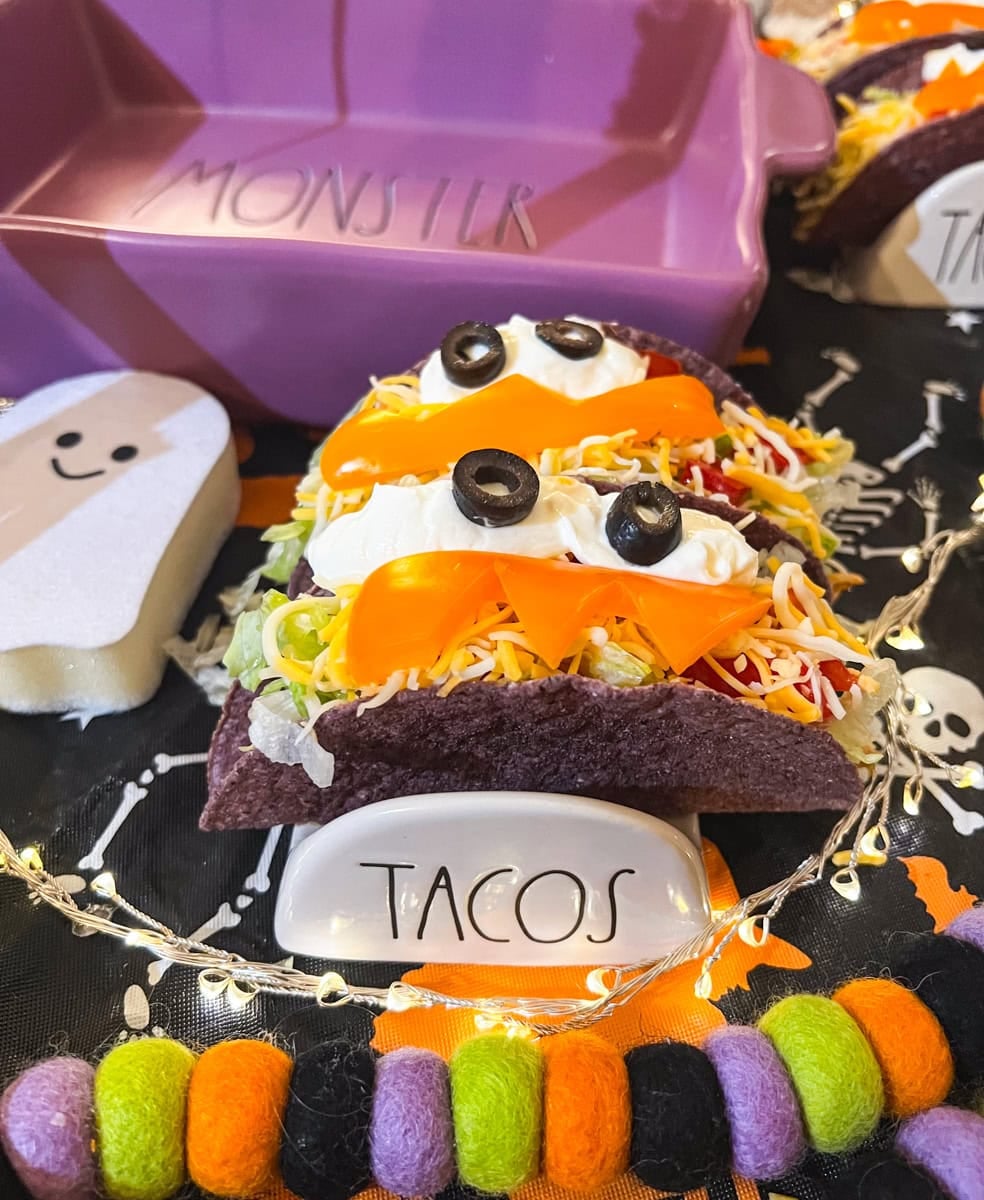 Two Halloween monster tacos in purple shells decorated with sour cream eyes, black olives, and orange bell pepper fangs, displayed with a Monster dish and taco holder.