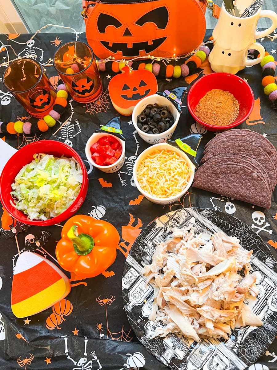 Overhead view of shredded rotisserie chicken, purple taco shells, lettuce, cherry tomatoes, shredded cheese, black olives, taco seasoning, and orange bell pepper arranged on a Halloween-themed tablecloth.