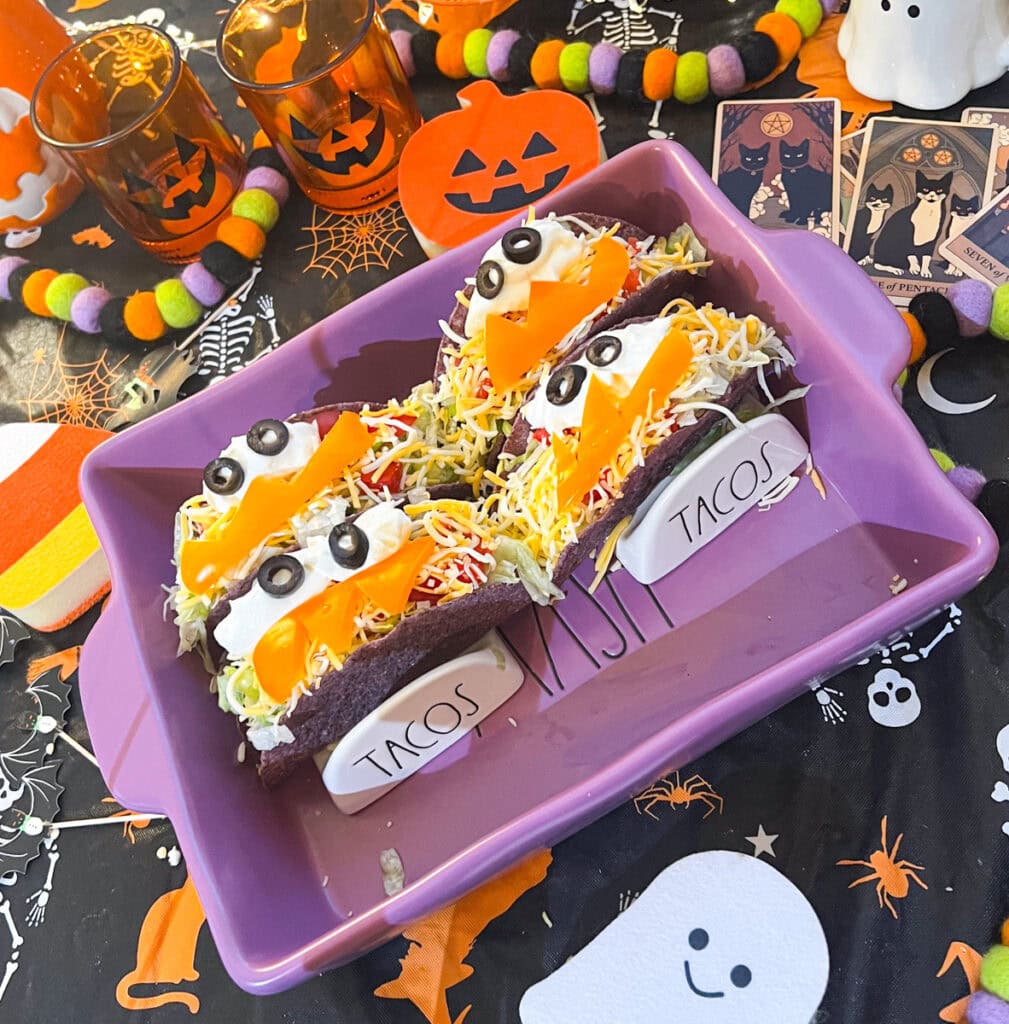 Three purple taco shells decorated with black olive eyes and orange bell pepper smiles with jagged teeth, arranged in a purple baking dish.