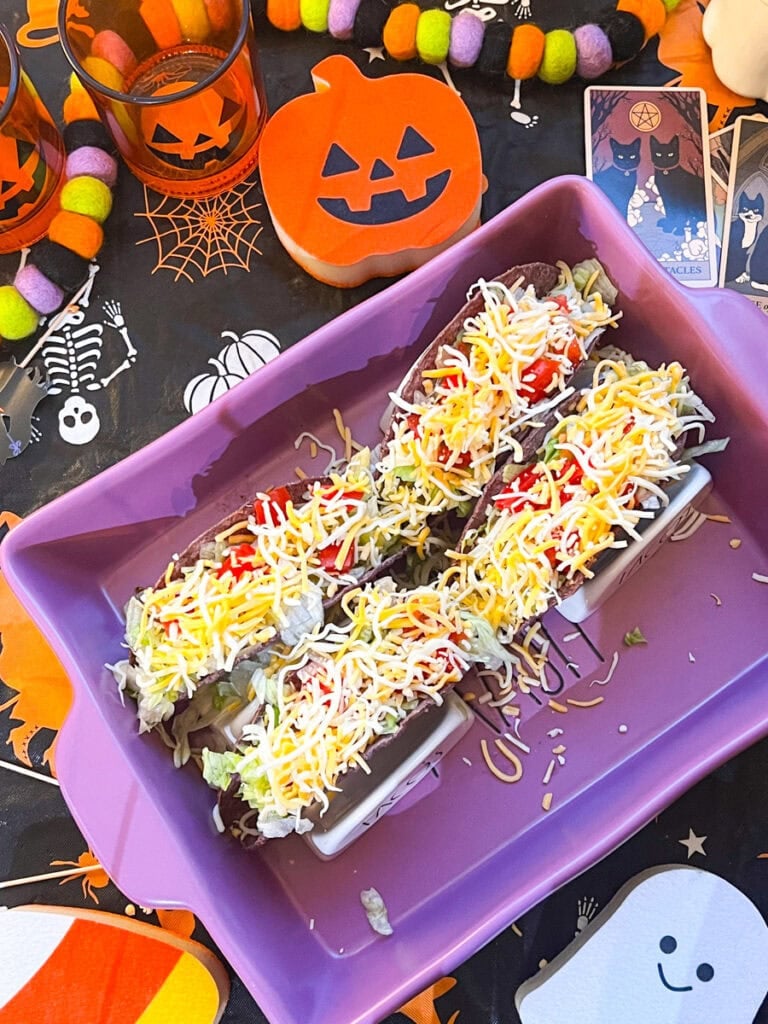 Three purple taco shells filled with shredded chicken, lettuce, shredded cheese, and diced cherry tomatoes in a purple baking dish.
