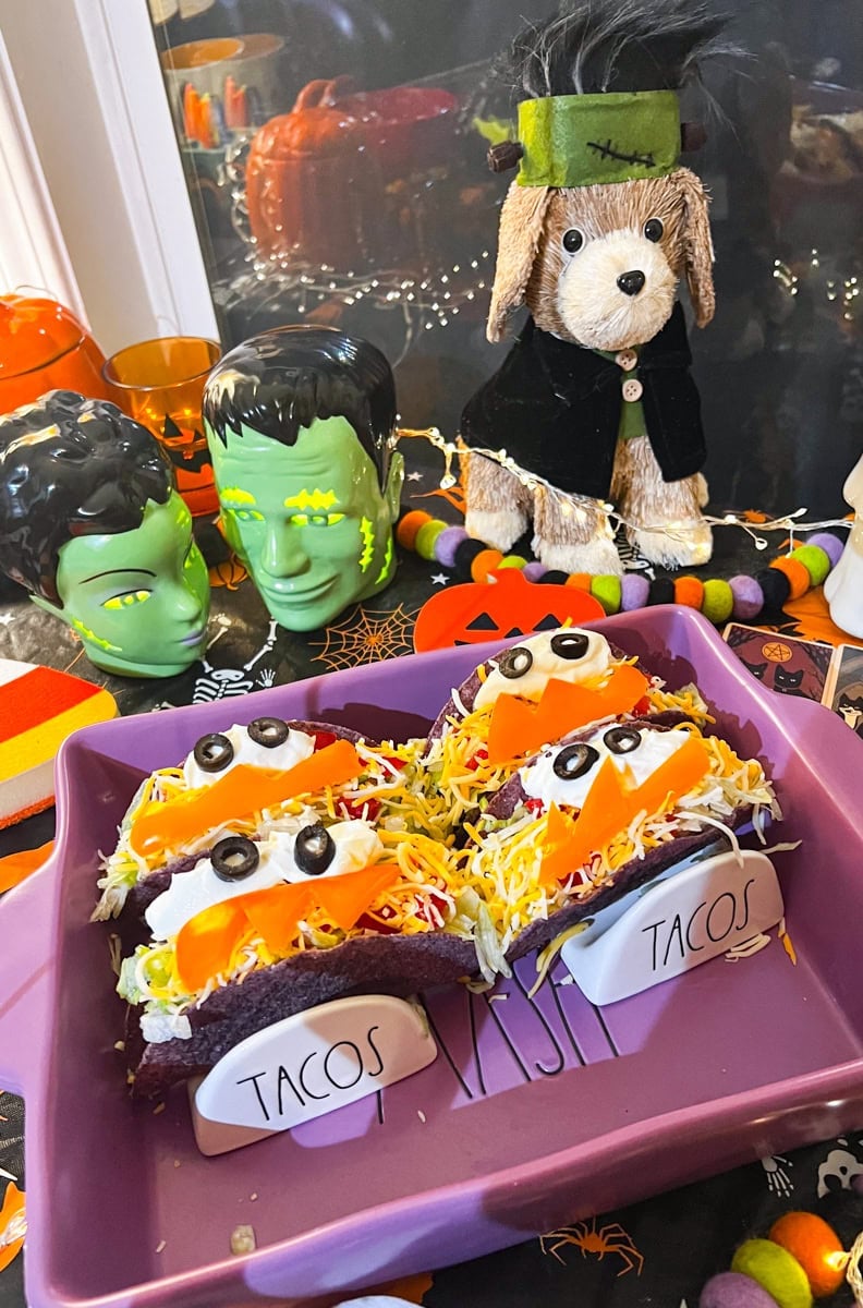 Three finished monster tacos with black olive eyes and orange bell pepper smiles served in a purple baking dish surrounded by spooky Halloween decorations.